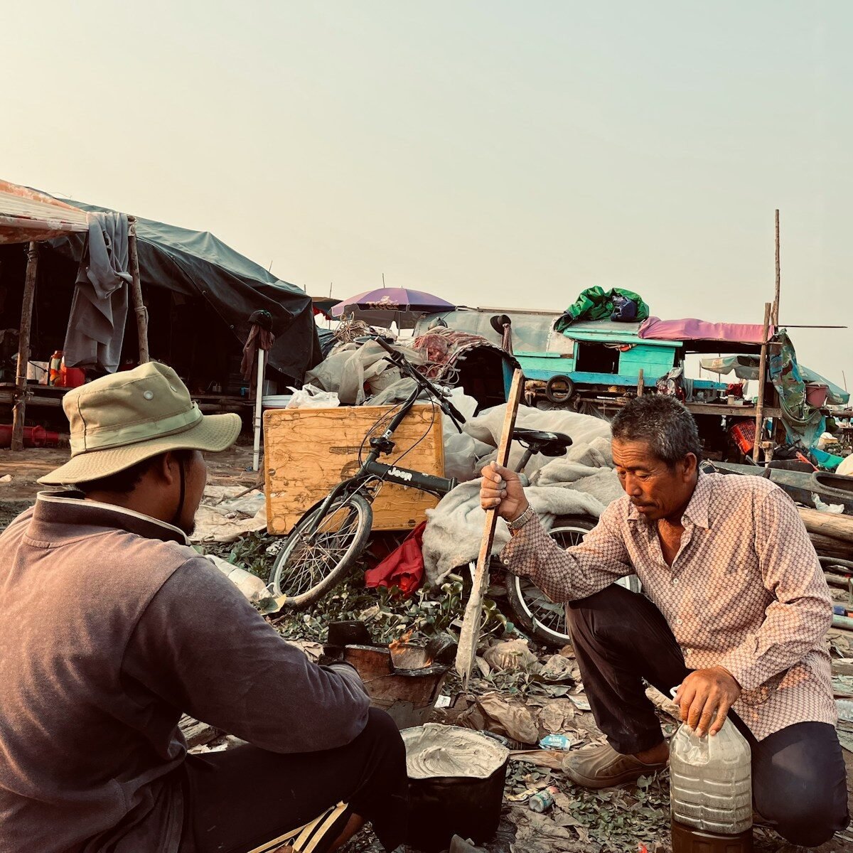 Two men cooking outdoors in a rustic settlement.